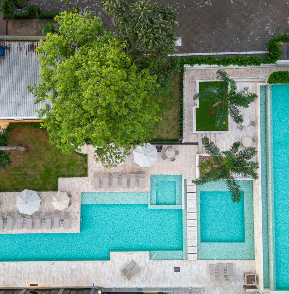 Vue aérienne d'une piscine moderne avec plusieurs bassins rectangulaires, entourée de chaises longues et de parasols, avec des arbres et un bâtiment à proximité.