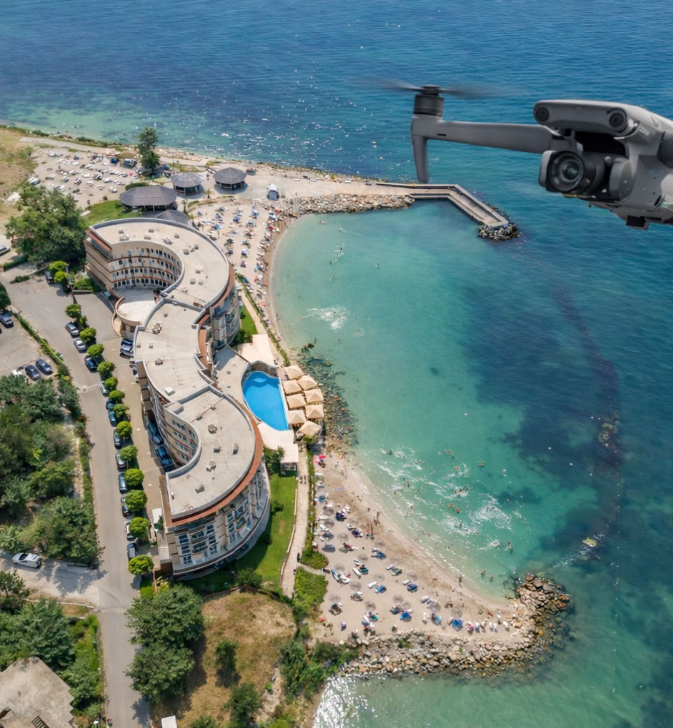 Vue aérienne d'un complexe hôtelier en bord de mer avec une piscine, une plage et un drone volant au-dessus de l'eau.